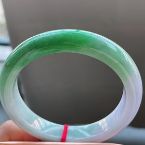 K&ouml;nnte beinhalten: Ein Jade-Armreif mit einem Farbverlauf von leuchtendem Gr&uuml;n zu sanftem Wei&szlig;. Die polierte Oberfl&auml;che reflektiert das Licht und zeigt die nat&uuml;rlichen Variationen des Steins. Der Armreif hat eine kreisf&ouml;rmige Form und scheint ein Schmuckst&uuml;ck zu sein. Durchmesser: 6.5 cm.
