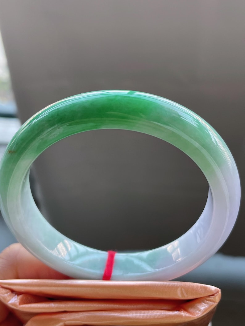 K&ouml;nnte beinhalten: Ein Jade-Armreif mit einem Farbverlauf von leuchtendem Gr&uuml;n zu sanftem Wei&szlig;. Die polierte Oberfl&auml;che reflektiert das Licht und zeigt die nat&uuml;rlichen Variationen des Steins. Der Armreif hat eine kreisf&ouml;rmige Form und scheint ein Schmuckst&uuml;ck zu sein. Durchmesser: 6.5 cm.