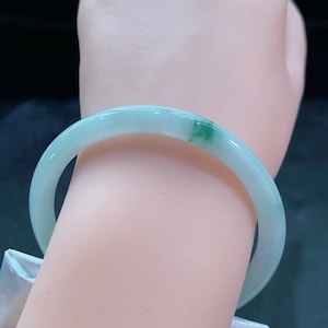 May include: A light green jade bangle bracelet. The bracelet has a smooth, polished surface with a translucent appearance. A small area of darker green is visible on the bracelet. The bangle is worn on a wrist.
