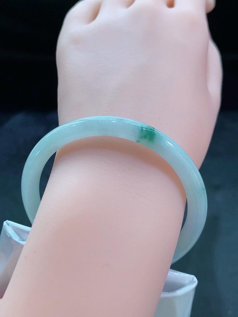 May include: A light green jade bangle bracelet. The bracelet has a smooth, polished surface with a translucent appearance. A small area of darker green is visible on the bracelet. The bangle is worn on a wrist.