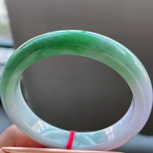 K&ouml;nnte beinhalten: Ein Jade-Armreif mit einem Farbverlauf von lebendigem Gr&uuml;n zu einem sanften, hellen Gr&uuml;n. Die polierte Oberfl&auml;che reflektiert das Licht und hebt die nat&uuml;rlichen Farb- und Texturvariationen hervor. Der Armreif hat eine kreisf&ouml;rmige Form.