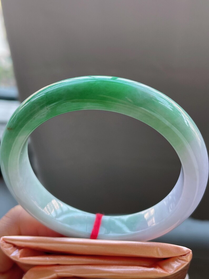 K&ouml;nnte beinhalten: Ein Jade-Armreif mit einem Farbverlauf von lebendigem Gr&uuml;n zu einem sanften, hellen Gr&uuml;n. Die polierte Oberfl&auml;che reflektiert das Licht und hebt die nat&uuml;rlichen Farb- und Texturvariationen hervor. Der Armreif hat eine kreisf&ouml;rmige Form.