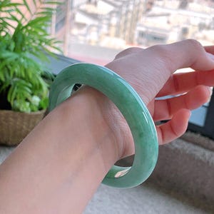 May include: A light green jadeite bangle bracelet. The smooth, polished surface reflects light, showcasing the natural variations in color. The jade bracelet is a classic piece of jewelry.