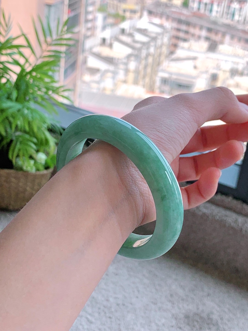 May include: A light green jadeite bangle bracelet. The smooth, polished surface reflects light, showcasing the natural variations in color. The jade bracelet is a classic piece of jewelry.