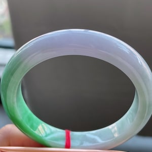 K&ouml;nnte beinhalten: Ein transluzentes Jade-Armreif-Armband. Das Armband wechselt von einem leuchtenden Gr&uuml;n zu einem sanften Lavendelton. Die glatte, polierte Oberfl&auml;che reflektiert das Licht und hebt die nat&uuml;rlichen Farb- und Texturvariationen hervor. Ein klassisches Schmuckst&uuml;ck.