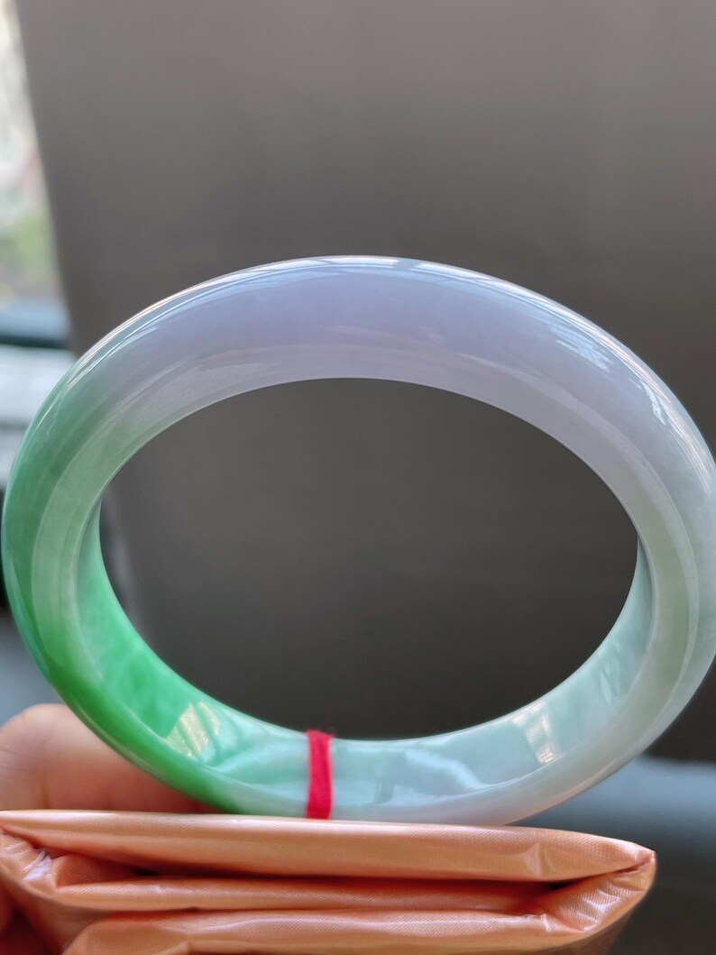 K&ouml;nnte beinhalten: Ein transluzentes Jade-Armreif-Armband. Das Armband wechselt von einem leuchtenden Gr&uuml;n zu einem sanften Lavendelton. Die glatte, polierte Oberfl&auml;che reflektiert das Licht und hebt die nat&uuml;rlichen Farb- und Texturvariationen hervor. Ein klassisches Schmuckst&uuml;ck.
