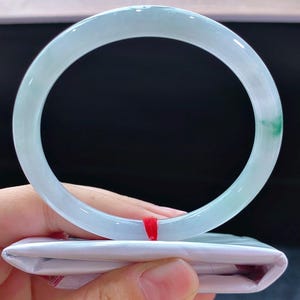 May include: A pale green jade bangle. The circular bracelet is translucent with a subtle colour variation and a hint of green. It is held above a white package with a red ribbon. The bracelet's diameter is approximately 6.5 cm.