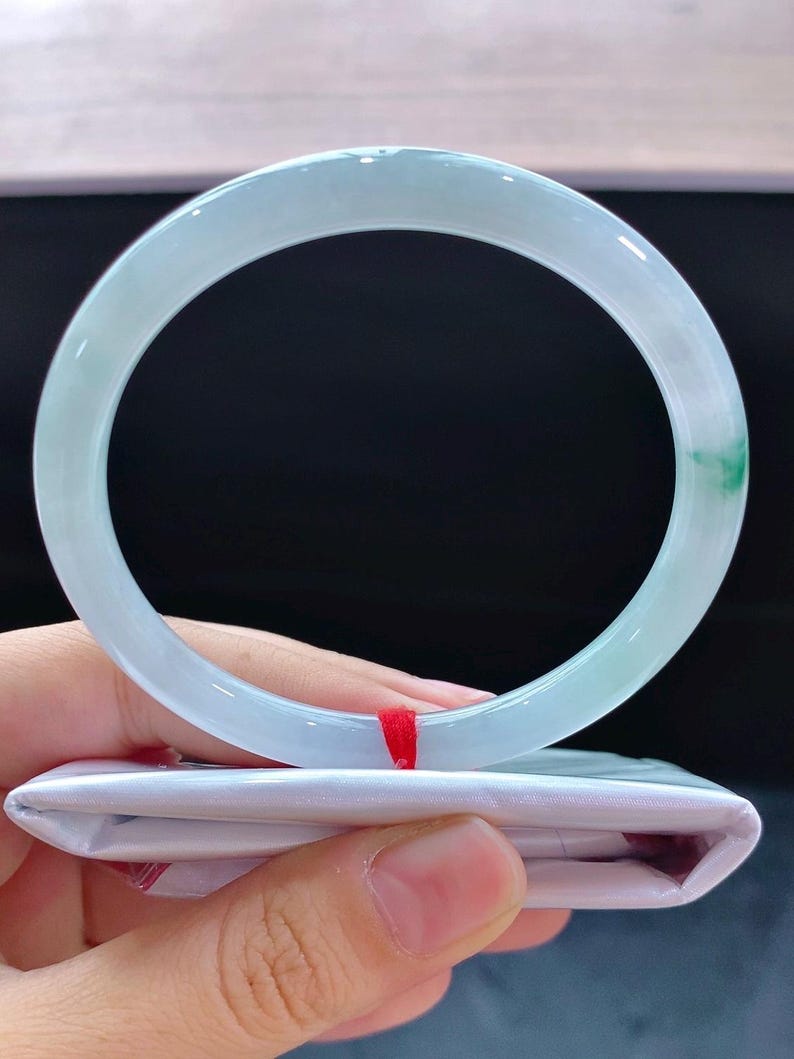 May include: A pale green jade bangle. The circular bracelet is translucent with a subtle colour variation and a hint of green. It is held above a white package with a red ribbon. The bracelet's diameter is approximately 6.5 cm.