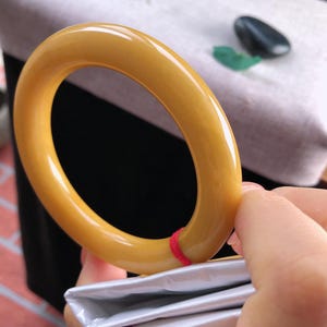 K&ouml;nnte beinhalten: Ein glattes, bernsteinfarbenes Armreif-Armband. Das kreisf&ouml;rmige Armband wird von einer Hand gehalten, wobei ein roter Faden sichtbar ist. Der Hintergrund umfasst eine wei&szlig;e Oberfl&auml;che und ein schwarzes Objekt.