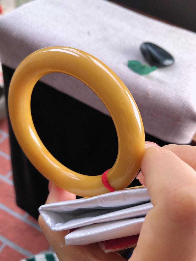 K&ouml;nnte beinhalten: Ein glattes, bernsteinfarbenes Armreif-Armband. Das kreisf&ouml;rmige Armband wird von einer Hand gehalten, wobei ein roter Faden sichtbar ist. Der Hintergrund umfasst eine wei&szlig;e Oberfl&auml;che und ein schwarzes Objekt.
