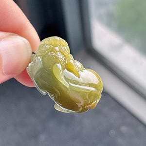 Peut inclure: Un pendentif en jade vert olive translucide sculpt&eacute; en forme d'une petite figurine stylis&eacute;e. Le jade pr&eacute;sente des zones de vert plus clair et plus fonc&eacute;, avec une surface lisse et polie. La figurine a un corps arrondi et des traits du visage d&eacute;taill&eacute;s.