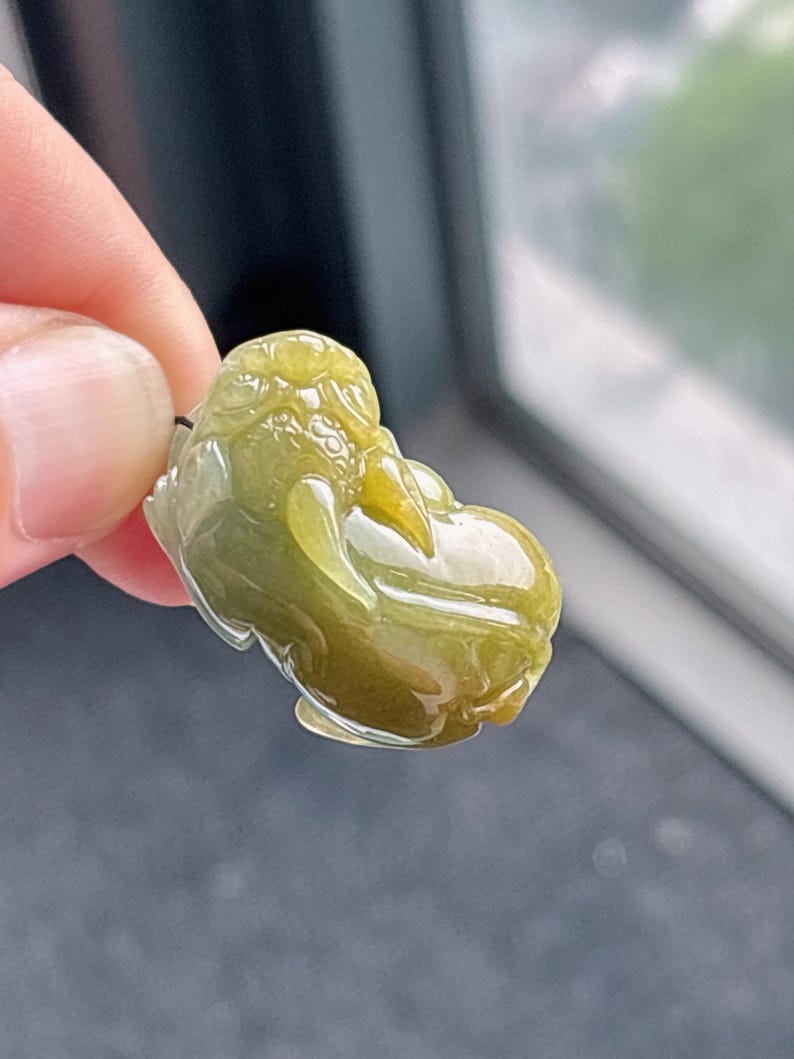 Peut inclure: Un pendentif en jade vert olive translucide sculpt&eacute; en forme d'une petite figurine stylis&eacute;e. Le jade pr&eacute;sente des zones de vert plus clair et plus fonc&eacute;, avec une surface lisse et polie. La figurine a un corps arrondi et des traits du visage d&eacute;taill&eacute;s.