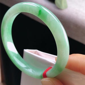 May include: A smooth, polished jade bangle bracelet featuring a translucent light green color with a distinct, vibrant emerald green patch. The circular jewelry piece has a rounded profile and is accented with a small, thin red string tied around one side.