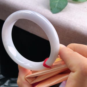 May include: A white jade bangle bracelet. The smooth, polished bracelet is held by a person's hand, revealing its circular form. The jade has a translucent quality, and a red string is attached to the bracelet. The background is blurred.