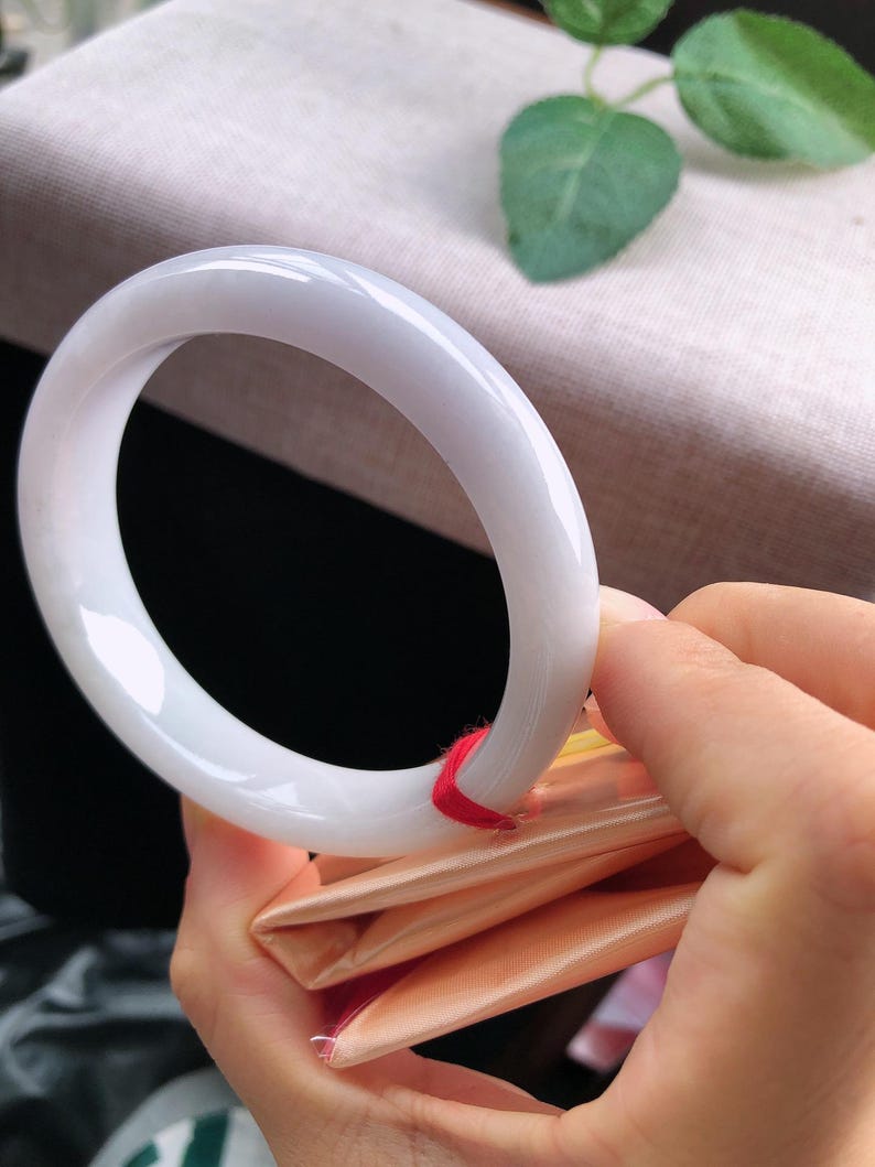 May include: A white jade bangle bracelet. The smooth, polished bracelet is held by a person's hand, revealing its circular form. The jade has a translucent quality, and a red string is attached to the bracelet. The background is blurred.