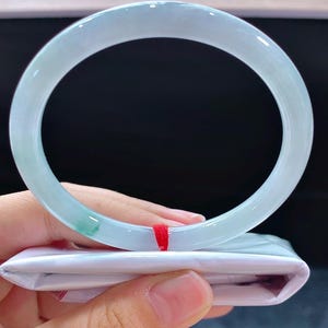 May include: A pale green jadeite bangle bracelet. The smooth, polished surface of the bracelet has a light, pastel green hue with subtle colour variations. The bracelet is held over a white package with a red ribbon.