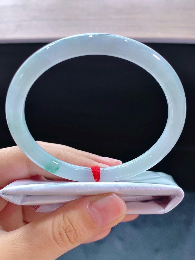 May include: A pale green jadeite bangle bracelet. The smooth, polished surface of the bracelet has a light, pastel green hue with subtle colour variations. The bracelet is held over a white package with a red ribbon.