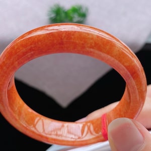 May include: A smooth, polished bangle bracelet crafted from translucent orange jadeite. The circular accessory features a vibrant, mottled reddish-orange hue with natural internal variations and a glossy, reflective surface finish throughout.