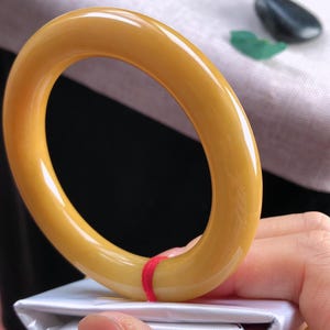 K&ouml;nnte beinhalten: Ein glattes, goldgelbes Armreif-Armband. Das Armband wird &uuml;ber einer wei&szlig;en Oberfl&auml;che gehalten, mit einem roten Faden gesichert. Der Armreif hat eine gl&auml;nzende Oberfl&auml;che und eine abgerundete Form. Ein kleiner, dunkler Stein und ein gr&uuml;nes Blatt sind im Hintergrund.