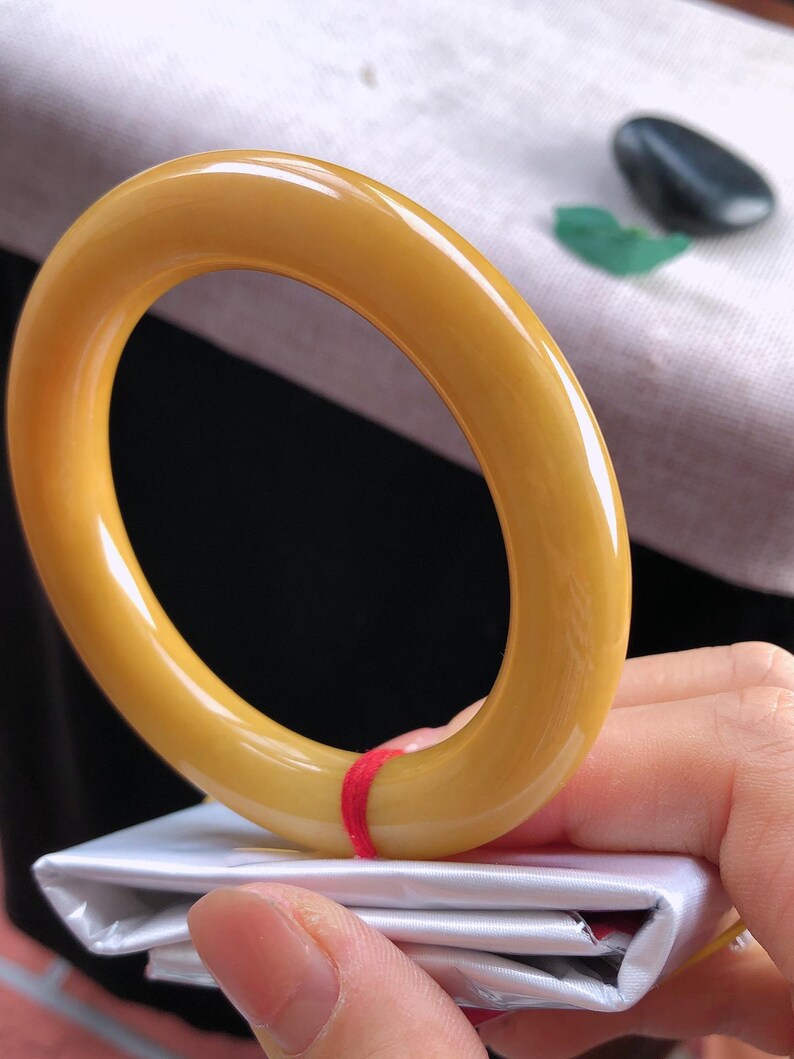 K&ouml;nnte beinhalten: Ein glattes, goldgelbes Armreif-Armband. Das Armband wird &uuml;ber einer wei&szlig;en Oberfl&auml;che gehalten, mit einem roten Faden gesichert. Der Armreif hat eine gl&auml;nzende Oberfl&auml;che und eine abgerundete Form. Ein kleiner, dunkler Stein und ein gr&uuml;nes Blatt sind im Hintergrund.