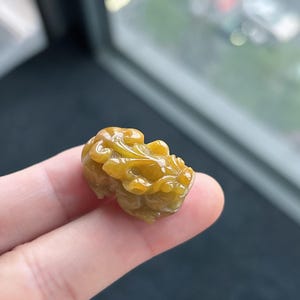 Pu&ograve; includere: Una statuetta di giada giallo dorato scolpita, tenuta tra le dita. La statuetta raffigura una creatura mitica con dettagli intricati, che mostra una superficie liscia e lucida. Il colore della giada varia dal giallo chiaro al giallo scuro.