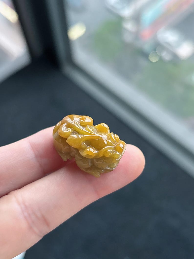 Pu&ograve; includere: Una statuetta di giada giallo dorato scolpita, tenuta tra le dita. La statuetta raffigura una creatura mitica con dettagli intricati, che mostra una superficie liscia e lucida. Il colore della giada varia dal giallo chiaro al giallo scuro.