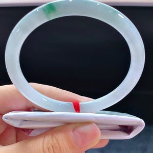 May include: A pale blue jade bangle with a hint of green. The circular bracelet is smooth and sits on folded white paper. A red string is visible.