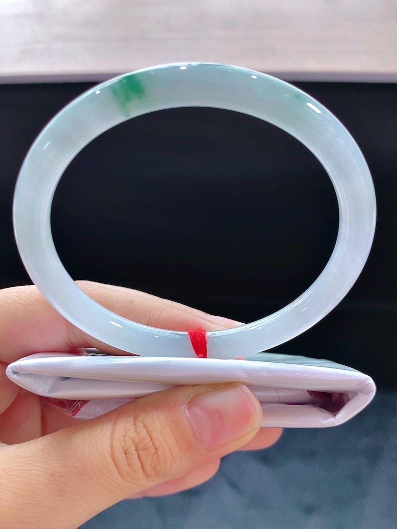 May include: A pale blue jade bangle with a hint of green. The circular bracelet is smooth and sits on folded white paper. A red string is visible.
