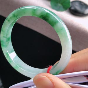 May include: A jade bangle bracelet, displaying a mix of light green and white tones. The polished surface reflects light, highlighting the natural colour variations. The bracelet is held by a hand, with a red string attached.