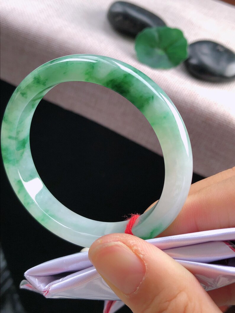 May include: A jade bangle bracelet, displaying a mix of light green and white tones. The polished surface reflects light, highlighting the natural colour variations. The bracelet is held by a hand, with a red string attached.