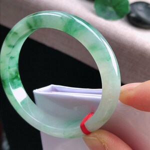 May include: A jade bangle bracelet, displaying a mix of light green and white tones. The polished surface reflects light, highlighting the natural colour variations. A red string is tied around the bracelet.