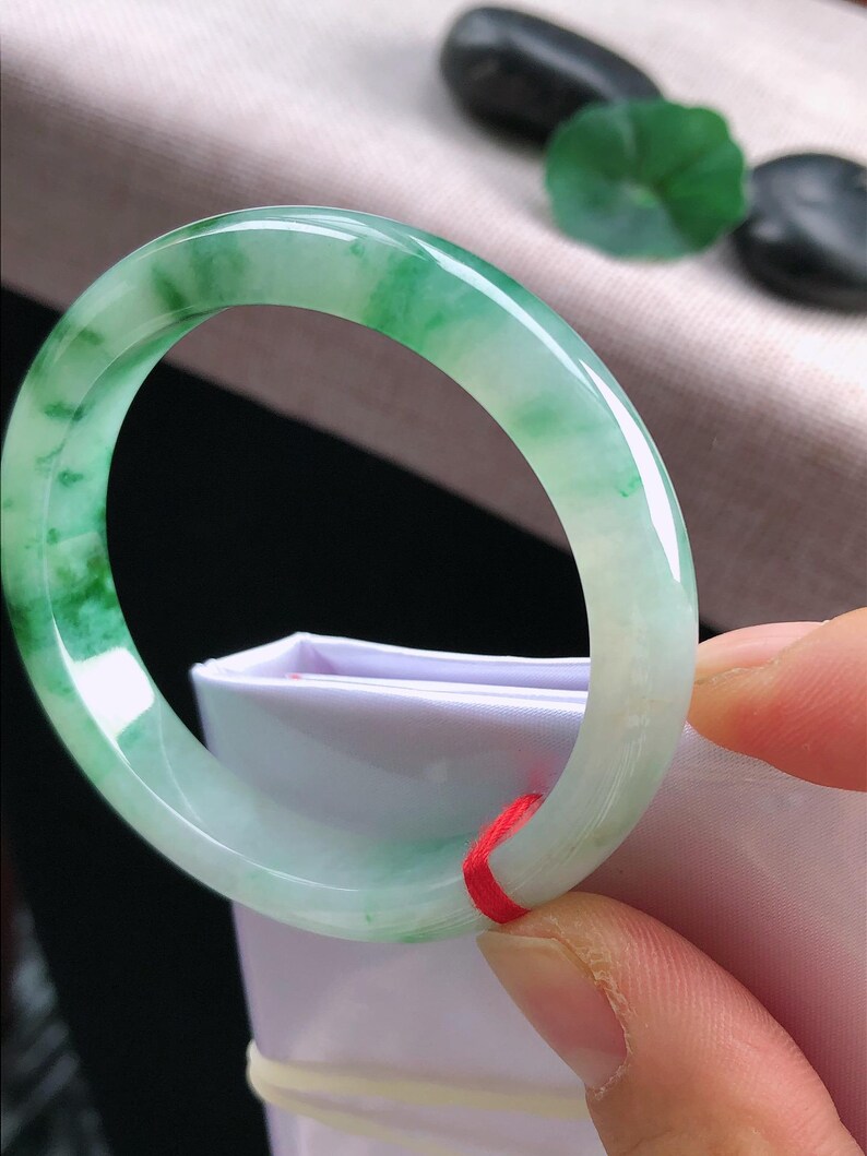 May include: A jade bangle bracelet, displaying a mix of light green and white tones. The polished surface reflects light, highlighting the natural colour variations. A red string is tied around the bracelet.