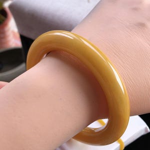 K&ouml;nnte beinhalten: Ein glattes, gelb-oranges Armreif-Armband. Das Armband ist ein massives Band aus poliertem Material mit abgerundeter Form. Die Farbe ist ein warmer, goldener Farbton. Das Armband wird am Handgelenk getragen.