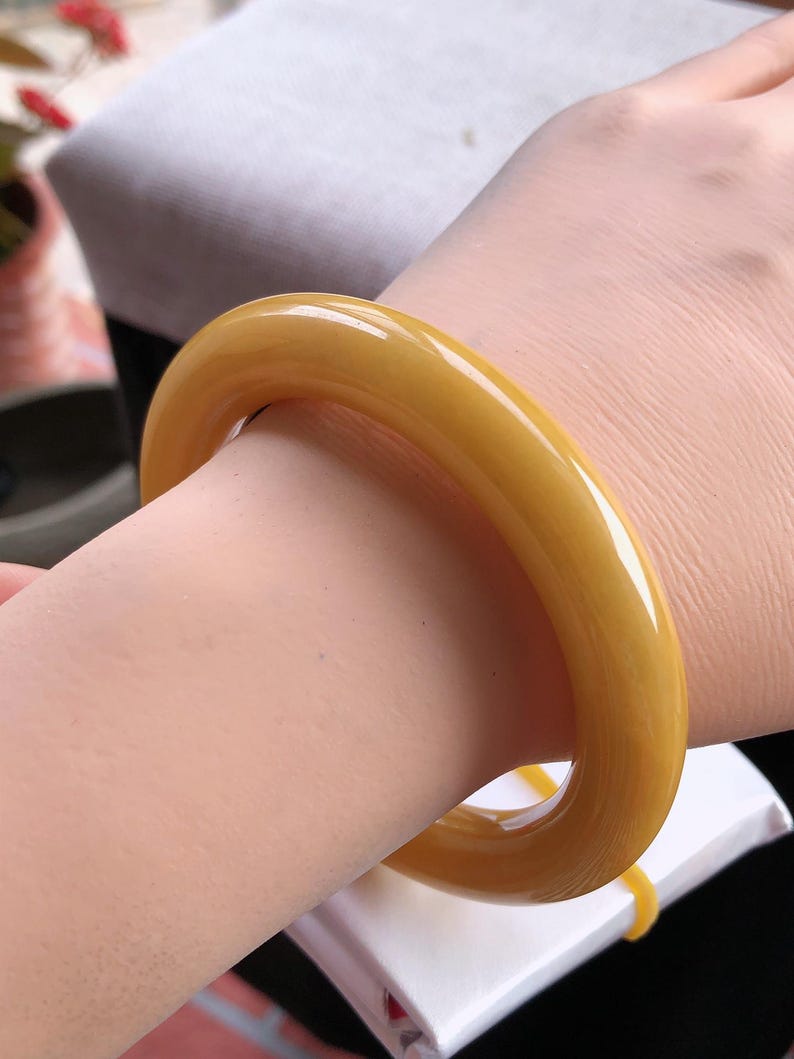 K&ouml;nnte beinhalten: Ein glattes, gelb-oranges Armreif-Armband. Das Armband ist ein massives Band aus poliertem Material mit abgerundeter Form. Die Farbe ist ein warmer, goldener Farbton. Das Armband wird am Handgelenk getragen.