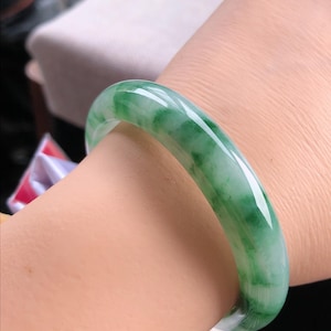 May include: A jade bangle bracelet with a smooth, polished surface. The bracelet features a translucent green colour with white marbling. The bangle is a circular shape and appears to be worn on a wrist.