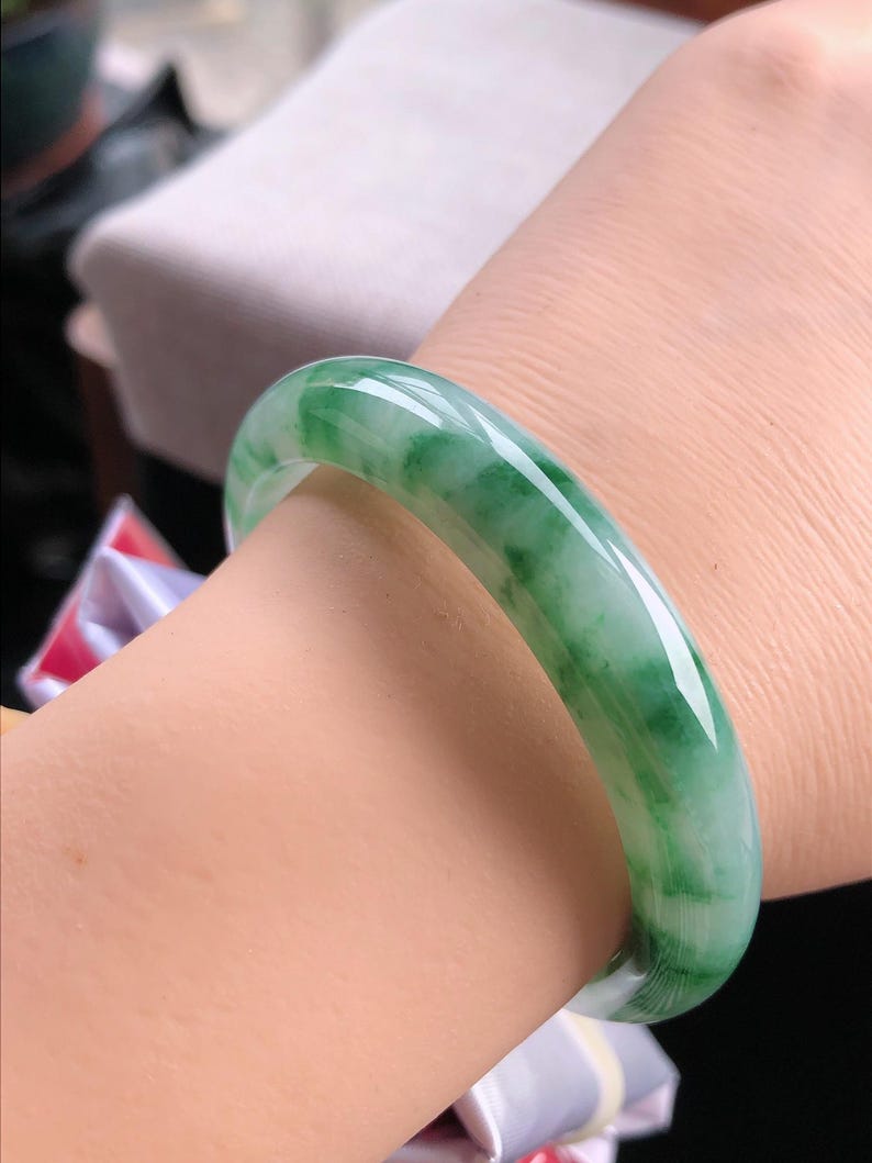 May include: A jade bangle bracelet with a smooth, polished surface. The bracelet features a translucent green colour with white marbling. The bangle is a circular shape and appears to be worn on a wrist.