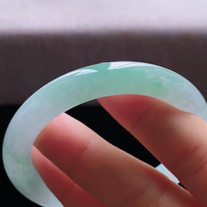 Op de afbeelding: Een doorschijnende jade armband. De jade heeft een lichtgroene kleur met een glad, gepolijst oppervlak. De armband heeft een cirkelvorm en lijkt een massief stuk jade te zijn. Het licht reflecteert op het oppervlak en benadrukt de kleurvariaties.