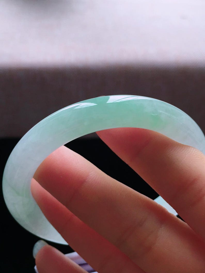 Op de afbeelding: Een doorschijnende jade armband. De jade heeft een lichtgroene kleur met een glad, gepolijst oppervlak. De armband heeft een cirkelvorm en lijkt een massief stuk jade te zijn. Het licht reflecteert op het oppervlak en benadrukt de kleurvariaties.