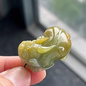 Peut inclure: Un pendentif en jade sculpt&eacute; en forme de cr&eacute;ature mythique. Le pendentif est un m&eacute;lange de teintes vertes et jaunes, avec des d&eacute;tails complexes. La sculpture est tenue par une ficelle noire.
