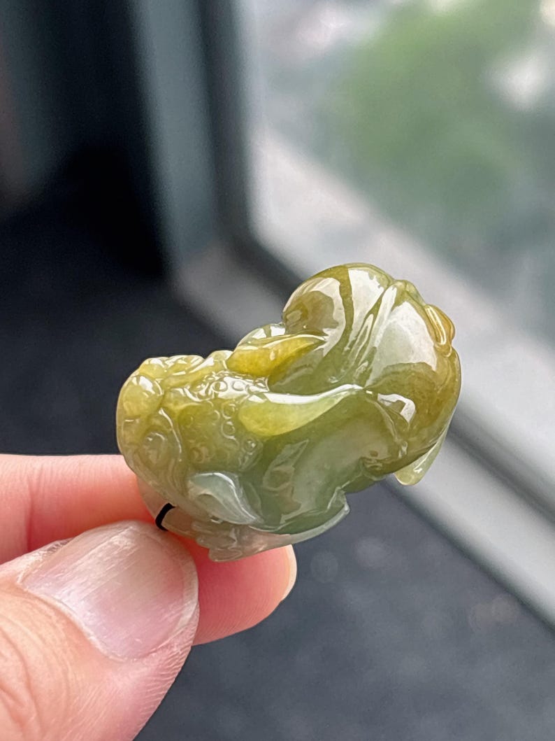 Peut inclure: Un pendentif en jade sculpt&eacute; en forme de cr&eacute;ature mythique. Le pendentif est un m&eacute;lange de teintes vertes et jaunes, avec des d&eacute;tails complexes. La sculpture est tenue par une ficelle noire.