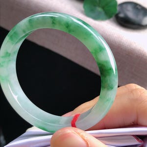May include: A jade bangle bracelet in shades of green and white. The bracelet has a smooth, polished finish and a circular design. The jade appears to be a natural stone, with a translucent quality.