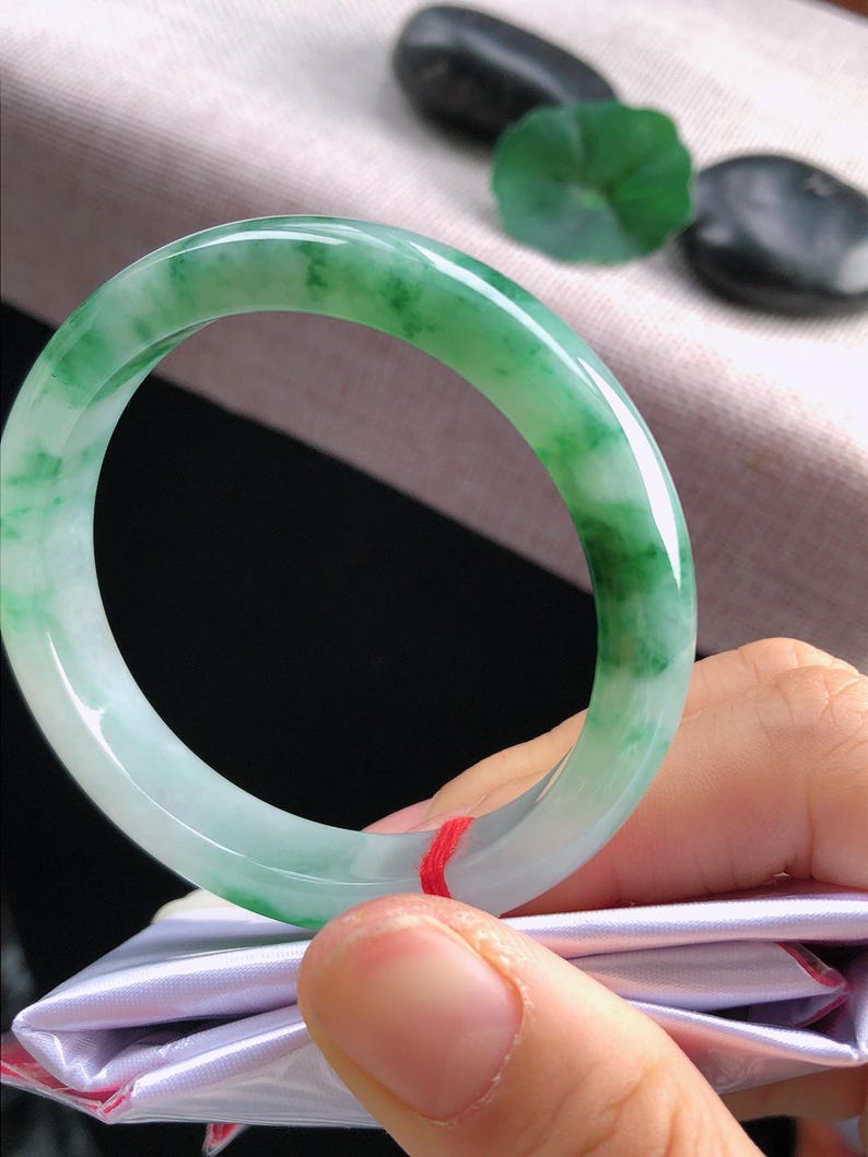 May include: A jade bangle bracelet in shades of green and white. The bracelet has a smooth, polished finish and a circular design. The jade appears to be a natural stone, with a translucent quality.