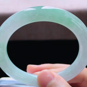 Op de afbeelding: Een lichtgroene jade armband. Het gepolijste oppervlak reflecteert licht en toont de natuurlijke kleurvariaties. De armband heeft een solide, cirkelvormig ontwerp, ideaal voor dagelijks gebruik. Diameter ongeveer 6,5 cm.