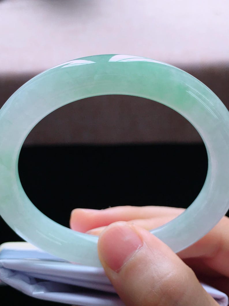 Op de afbeelding: Een lichtgroene jade armband. Het gepolijste oppervlak reflecteert licht en toont de natuurlijke kleurvariaties. De armband heeft een solide, cirkelvormig ontwerp, ideaal voor dagelijks gebruik. Diameter ongeveer 6,5 cm.
