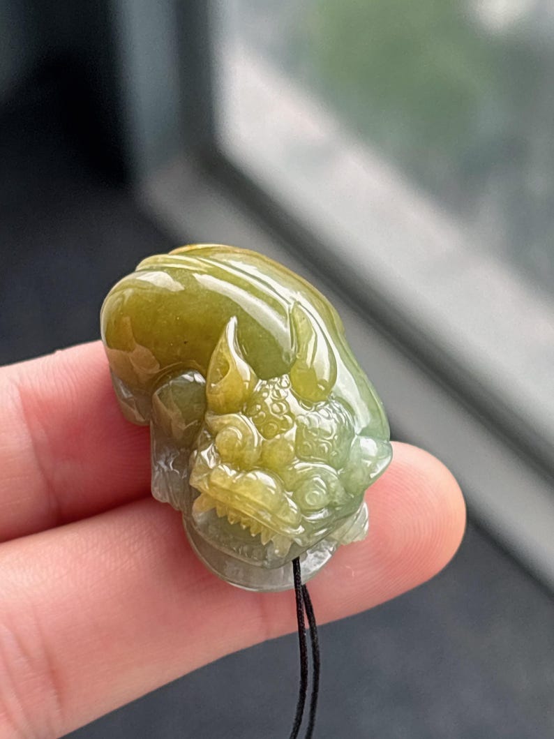 Peut inclure: Pendentif en jade sculpt&eacute; en forme de cr&eacute;ature mythique. Le pendentif est un m&eacute;lange de teintes vertes et jaunes, avec des d&eacute;tails complexes. Un cordon noir est attach&eacute; pour le porter.
