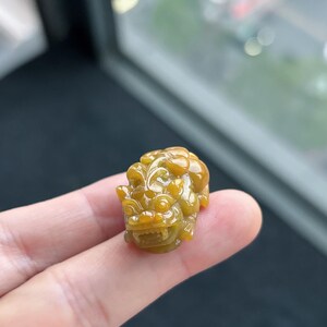 Pu&ograve; includere: Piccola statuetta in giada scolpita, nei toni del giallo e del marrone. La scultura raffigura una creatura mitica con dettagli fini, tenuta tra le dita. La giada ha una finitura lucida e lucente, e l'intaglio mette in risalto l'abilit&agrave; dell'artista.