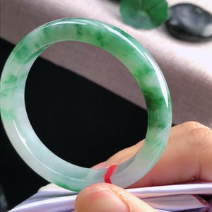 May include: A jade bangle bracelet, displaying a mix of light green and white tones. The polished surface reflects light, highlighting the natural colour variations. The bracelet is held by a person.