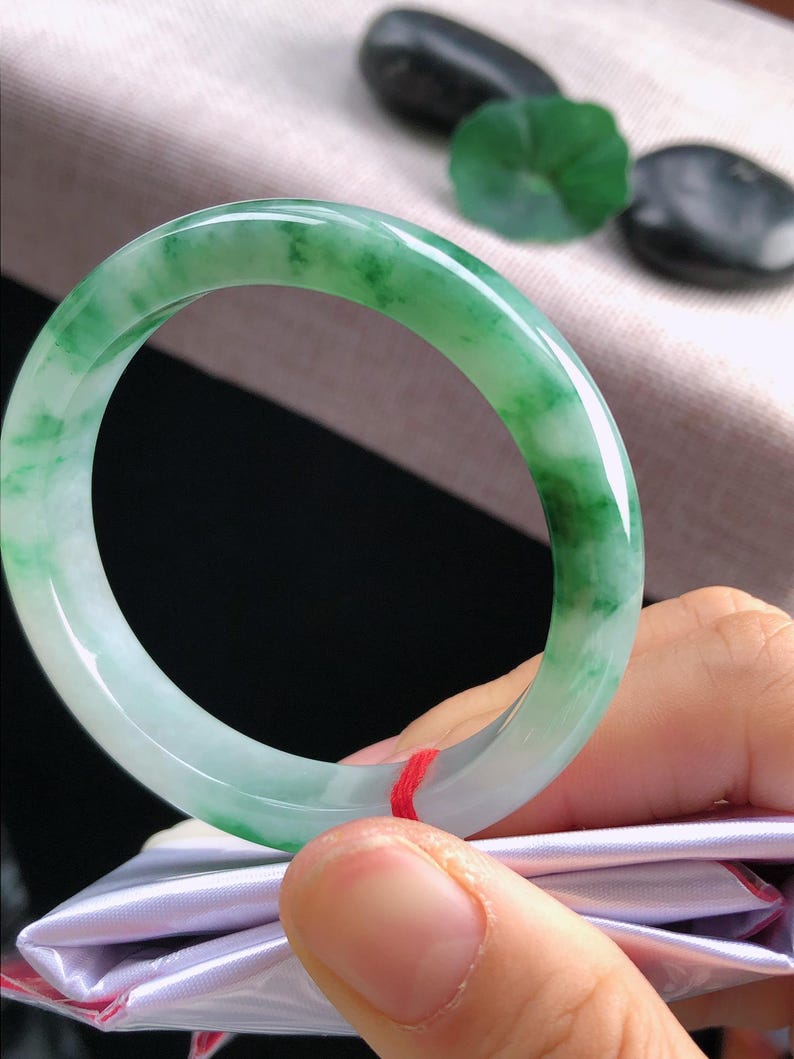 May include: A jade bangle bracelet, displaying a mix of light green and white tones. The polished surface reflects light, highlighting the natural colour variations. The bracelet is held by a person.
