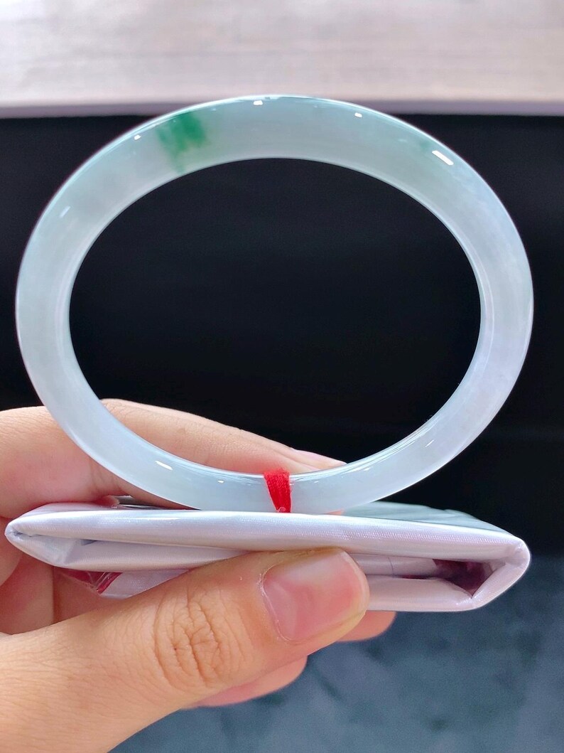 May include: A translucent jade bangle bracelet with a pale green and white colour gradient. The jade is smooth and polished, with a touch of vibrant green. The bracelet is held over a white folded package with a red ribbon.