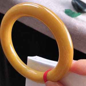 K&ouml;nnte beinhalten: Ein poliertes, bernsteinfarbenes Armreif-Armband. Das kreisf&ouml;rmige Armband wird von einer Hand gehalten. Das Armband hat eine glatte, gl&auml;nzende Oberfl&auml;che. Der Hintergrund ist verschwommen, mit Andeutungen von Wei&szlig; und Schwarz.